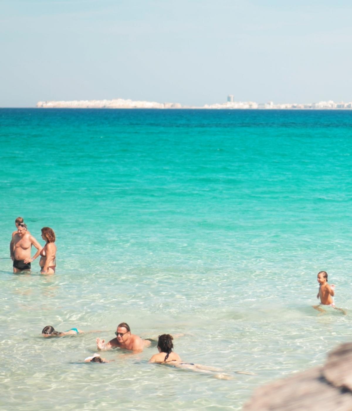 People swimming in clear sea with city in background.